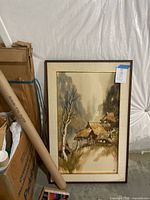 Framed watercolor painting of thatched roof dwellings on grassy slope with trees and signed lower right.