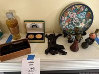 Full overview of the lot showing all items on a white mantel including the Leatherman tool in box, decorative plate on black stand with floral and dragon motifs, two small vases, wooden sundial carving, and bottle of ginseng root liquor.