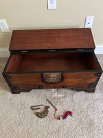 Open Asian wooden trunk on beige carpet with decorative brass lock plate on front. The accompanying decorative key and matching tassel lay nearby on the carpet.
