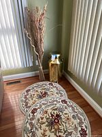 Photo showing two small round floral rugs on hardwood floor, next to a large shiny brass jug and a dried wood decorative arrangement.
