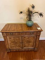 Front view of Century wooden buffet with drawer and double doors, wood grain finish, wheels visible below. Small Cerulean vase with peacock feathers on top.