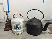 Photo showing the cast iron kettle, white Asian porcelain water jug, and cast-iron deer doorstop side by side on a concrete floor.