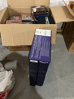 Box filled with assorted CDs and two blue hardcover books in slipcase lying on floor.