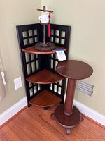 Full view of black and brown corner shelf unit with three triangular shelves, adjacent round wooden plant stand, and decorative Asian item with teacup on top shelf