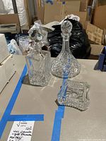 Two glass decanters and a glass ashtray sitting on a table in a basement room with boxes in the background.