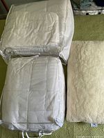 Photo showing full size quilted duvet and queen size polyester duvet in bags, alongside a merino wool covered pillow on green surface.