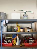 Shelf with garden pesticide containers, large ceramic flower pot, handheld garden sprinkler, bird feeder, and red bins labeled for extension cords and garden tools.