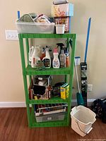 Full view of green plastic storage shelf with cleaning products on each tier and cleaning tools placed beside it.