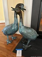 Pair of two metal decorative geese sculptures in cerulean and bronze tones, standing on hardwood floor near a doorway.