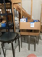 Four metal patio tables with clear glass tops, two dark brown and two medium gray, along with a cardboard box of light bulbs on top of one table. Image shows entire lot contents and condition.