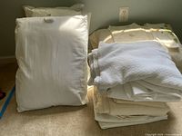 Photo showing 5 standard white pillows and folded assorted light-colored bedding items including a white cotton blanket.