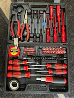 Top view of red and black screwdriver and plier tool set in black molded plastic case, including tape measure, socket bits, and electrical tape.