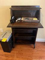 Small wooden secretary bookcase with drop down desk, with stapler, hole punch, calculator and other desk items on desktop, nonworking shredder beside it.