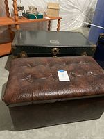 Shows steamer trunk and storage ottoman side by side on a concrete floor. Ottoman has a brown tufted top, trunk is black with metal fittings.