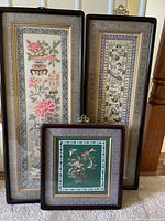 Three framed embroidered art pieces grouped together on floor for display. Includes two vertical rectangular panels and one smaller square panel.