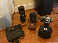 Four visible cameras (two folding bellows models, Rolleicord TLR, Nikomat SLR) and a pouch on hardwood floor.