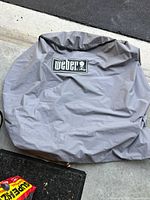 Gray Weber grill cover laid on the ground, showing the brand logo prominently.