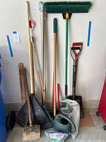 Photo of assorted yard and garden tools including shovels, rakes, a large wooden mallet, watering can, broom, and some poles leaning against a wall.