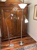 Two metal floor lamps placed on wooden floor in front of a carved wooden armoire. Left lamp has adjustable arm and square base. Right lamp is taller with round base, large frosted glass shade on top, and smaller frosted glass reading light attached to side.