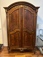 Full front view of cherry wood antique armoire showing detailed carved top and panel doors with gold floral decorations.