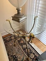 Angle view of glass top side table showing clear round glass surface and twisted metal base with brass accents, placed on patterned rug near window with white blinds.