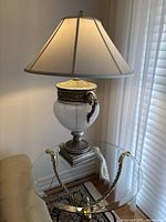 Glass table lamp with ornate bronze handles and beige fabric shade on a glass and brass table, near window blinds.