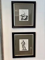 Two framed black and white prints hung on a white wall, one depicting an archer with a bow and the other a standing man.