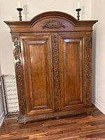 Front view of antique wooden armoire showing carved floral scrollwork, shell motif on top arch, and raised panel doors.