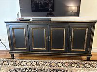 Front view of the black wooden TV stand with four cabinet doors and decorative metal handles under a mounted TV, sitting on patterned carpet.