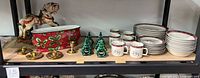 Wide view of shelf displaying Christmas-themed tableware and decor including brass candle holders, ceramic sleigh planter, ceramic Christmas tree and horse-drawn sleigh figurines.