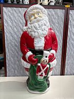 Front view of large plastic Santa Claus yard ornament, showing detailed facial features, red suit, and green sack with toy soldier decoration.