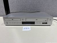 Front view of silver Samsung DVD-V5500 dual deck player with DVD and VHS slots, control buttons, display screen, and AV inputs.