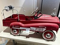 Side view showing red firetruck pedal car marked N. 287, with white metal frame at rear for ladder (missing), decal text, and rubber tires with white rims.