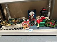 Shelf display showing Christmas Carol Clock box, holiday cookie platter, Santa figurines, gingerbread house decoration, black lantern, and Christmas wreath.