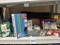 Shelf displaying a 150w floodlight box, battery free crank lantern box, assorted small hardware items like tacks, command strips, felt pads, and a flexible black hose coiled at the back, along with small plastic storage crate.