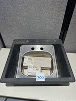 Photo showing stainless steel sink with three holes on back ledge and a rounded front edge, resting inside the black kitchen sink. Boxes and label number 303 placed inside the sink.