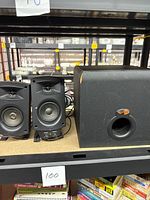 Two black Klipsch speakers and one Klipsch subwoofer on a shelf. Speakers have visible midrange drivers and tweeters, subwoofer shows surface wear.