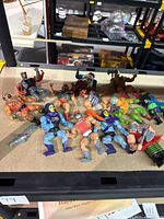 Twelve vintage Masters of the Universe action figures displayed lying down in a group, showing a mix of characters with some holding weapons. Figures have varied colors and some wear visible.