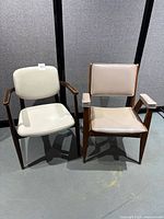 Two vintage mid-century modern armchairs side by side. Both have wooden frames and vinyl padded seats and backs. Visible wear on both, with scratches and peeling finish on one chair.