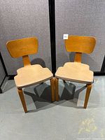 Pair of vintage Thonet chairs facing front showing wooden frames, beige padded seats, and curved backs, placed on concrete floor against gray backdrop.