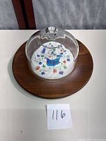 Photo showing the entire cheese plate set with wooden base, ceramic insert with blue bird and flowers, and glass dome lid on top.
