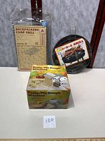 Lot contents on white surface showing all three items: boxed pasta machine, packaged camp grill, and round barbecue grill pan with handle.