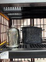 Black speckled enamel stock pot with lid and stainless steel percolator on shelf.
