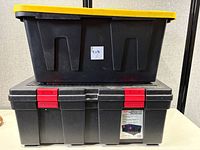 Black XXL Commander plastic bin with yellow lid stacked on top of black storage locker with red latches on white table.