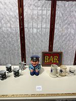 Plastic Cookie Cop jar in police uniform, three Toll Gate Class stoneware mugs, four black and white floral coffee mugs, cream mustache mug, and glitter art bar sign