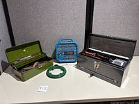 Photo showing two metal toolboxes, one green and one gray, along with a blue screwdriver bit case and a green fish tape reel on a white surface