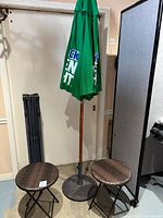Full view of green Bud Light Rock Green Light umbrella with wooden pole, weighted metal base, and two small brown woven collapsible tables/stools on either side, with folded collapsible canopy frame beside them.