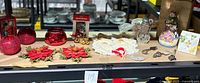 A collection of assorted Christmas decor on a table including four red glass votive holders, two red fabric poinsettia candle holders, a snowman snow globe, Swarovski crystal ornament, lace doily, and three silver-tone cheese cutters.
