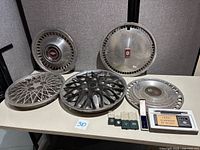 Six automotive hubcaps displayed on table with several VW and Audi pins and VW stick pin in packaging.