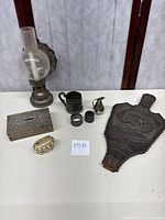 Photo showing all items arranged on a table including oil lamp with chimney, silver plated jewelry box, table lighter, two silver plate napkin rings, embossed decorative bellow, pewter creamer, and engraved silver plated cup.